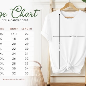 May include: A white t-shirt displayed on a wooden hanger, with a size chart to the left. The chart lists sizes XS to 5XL, with width and length measurements in inches. The text "Size Chart" is at the top, with "BELLA CANVAS 3001" below it.