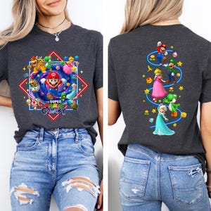 May include: Dark gray t-shirt featuring a colorful graphic of Super Mario characters, including Mario, Princess Peach, and Yoshi, with the text "The Super Mario Galaxy." The back of the shirt displays additional characters. The shirt is paired with blue jeans.