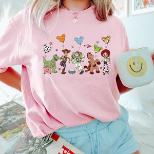Pu&ograve; includere: Una maglietta rosa chiaro con una grafica colorata dei personaggi di Toy Story. Il design include Woody, Buzz Lightyear e altri personaggi con palloncini. La maglietta &egrave; a girocollo e ha maniche corte.