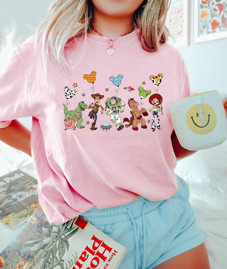 Pu&ograve; includere: Una maglietta rosa chiaro con una grafica colorata dei personaggi di Toy Story. Il design include Woody, Buzz Lightyear e altri personaggi con palloncini. La maglietta &egrave; a girocollo e ha maniche corte.