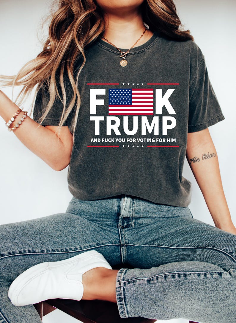 May include: Dark gray t-shirt with the text "F**k Trump" in white, featuring an American flag design. The shirt also includes the phrase "And F**k You For Voting For Him" in white. The model is wearing blue jeans and white shoes.