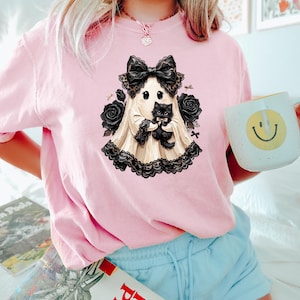 May include: A light pink t-shirt featuring a graphic of a cream-colored ghost wearing a large black lace bow and holding a small black kitten. The ghost is surrounded by black roses and black lace trim, creating a detailed, layered illustration.