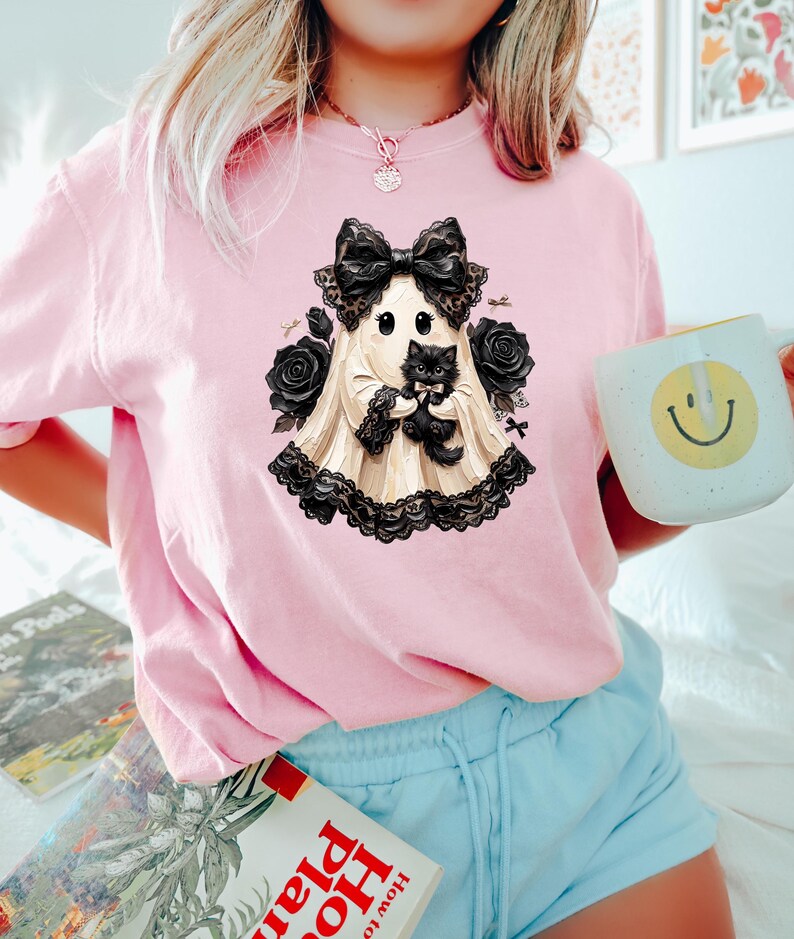 May include: A light pink t-shirt featuring a graphic of a cream-colored ghost wearing a large black lace bow and holding a small black kitten. The ghost is surrounded by black roses and black lace trim, creating a detailed, layered illustration.