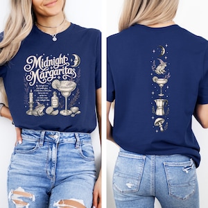 May include: A navy blue t-shirt featuring a front graphic with the text Midnight Margaritas, a margarita glass, a frog in a hat, a candle, and a bottle. The back graphic displays a vertical arrangement of a crescent moon, a frog, a margarita, a coffee pot, and a mushroom.