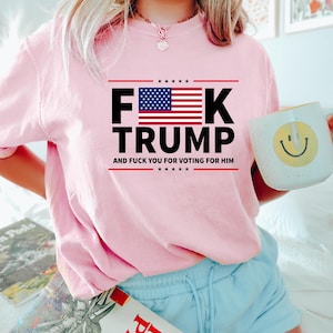 May include: A light pink sweatshirt with the text "F**K TRUMP" in black and red, with an American flag graphic. The text "AND F**K YOU FOR VOTING FOR HIM" is below. The person is holding a white mug with a yellow smiley face.