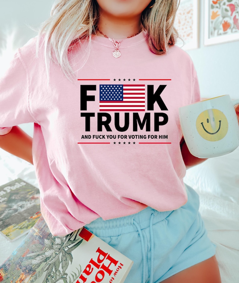 May include: A light pink sweatshirt with the text "F**K TRUMP" in black and red, with an American flag graphic. The text "AND F**K YOU FOR VOTING FOR HIM" is below. The person is holding a white mug with a yellow smiley face.