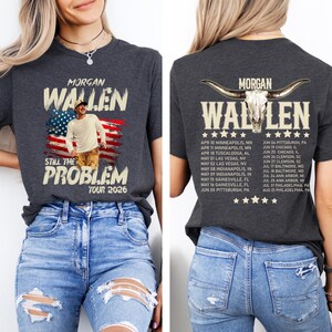 May include: Dark gray t-shirt featuring the text "MORGAN WALLEN" above an image of the American flag and a man. The shirt also includes the text "STILL THE PROBLEM TOUR 2026". The back of the shirt has a list of tour dates.