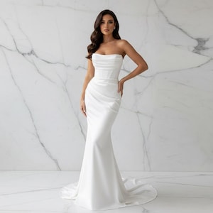 May include: A strapless white satin wedding gown with a fitted bodice and a mermaid silhouette. The dress features elegant draping and a flowing skirt. The model is standing in front of a marble wall.