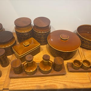 May include: A selection of brown ceramic kitchenware with wooden lids and trim. The set includes canisters, a rectangular box, a covered casserole dish, a bowl, small cups, and bottles, all with a repeating floral pattern.