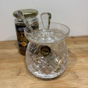 May include: A cut-glass sugar bowl with a lid and a small spoon, alongside a jar of honey. The sugar bowl features a diamond pattern and a clear handle. The honey jar has a gold lid and a label with text.