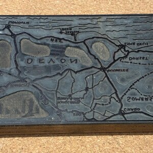 May include: A dark grey map of Devon, England, with etched lines and place names. The map includes names such as 'DEON', 'DOUZEL', and 'POWERSZEL'. The map is set in a wooden frame.
