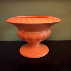 May include: An orange ceramic urn with a pedestal base. The urn has a wide, fluted rim and a detailed, curved body. The base is round and the colour is a solid, vibrant orange. The urn is likely for decorative purposes.