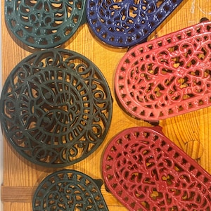 May include: A selection of decorative cast iron trivets in various colours, including green, blue, and red. The trivets feature intricate, openwork designs and are arranged on a wooden surface. The shapes include oval and round.