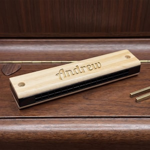 Puede incluir: Una armónica de madera clara con el nombre "Andrew" grabado. La armónica tiene detalles negros y está sobre una superficie de madera oscura. También se ven una púa y diapasones.