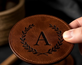 May include: A round, brown leather coaster with a stitched edge. The coaster features a black laurel wreath design around a large letter "A". The coaster is held by a hand, with a dark background.