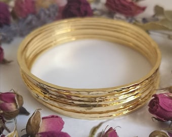 May include: A stack of six gold-colored bangles with a hammered texture. The bangles are arranged on a white surface, surrounded by dried pink roses and other dried flowers. The bangles have a shiny finish.