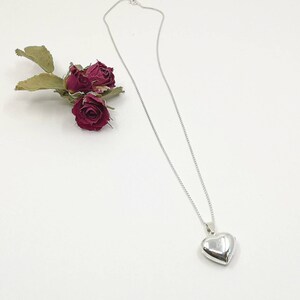 A sterling silver puffy heart locket on a silver chain. This heart necklace is on a white background with three dried red rose buds.