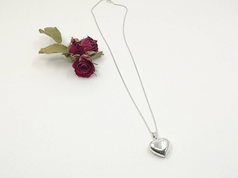 A sterling silver puffy heart locket on a silver chain. This heart necklace is on a white background with three dried red rose buds.