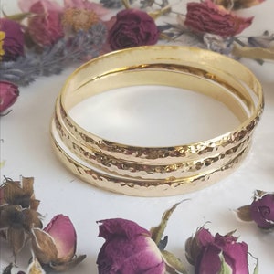 May include: Three gold-toned hammered bangle bracelets stacked on top of each other. The bracelets are smooth and shiny.