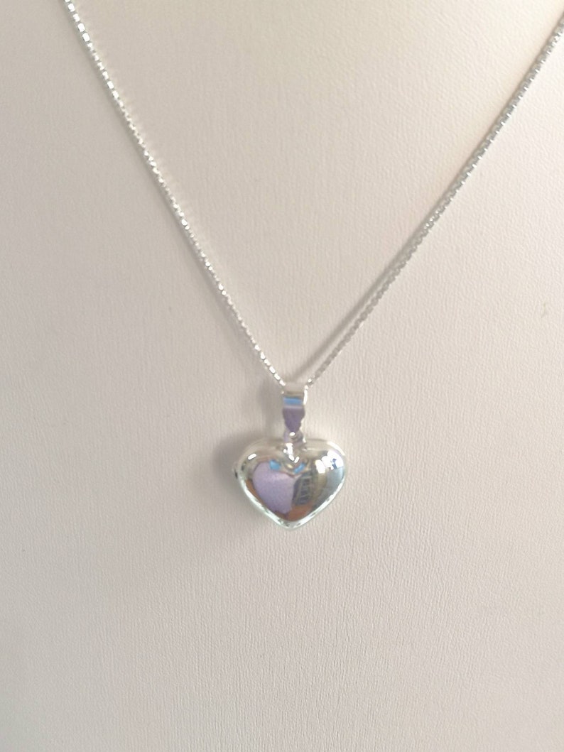 A sterling silver puffy heart locket on a silver chain. This heart necklace is on a white background. The locket is shiny and silver.