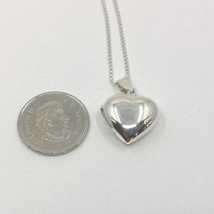 A sterling silver puffy heart locket on a silver chain. This heart necklace is on a white background next to a Canadian dime to show the size and scale of the pendant.