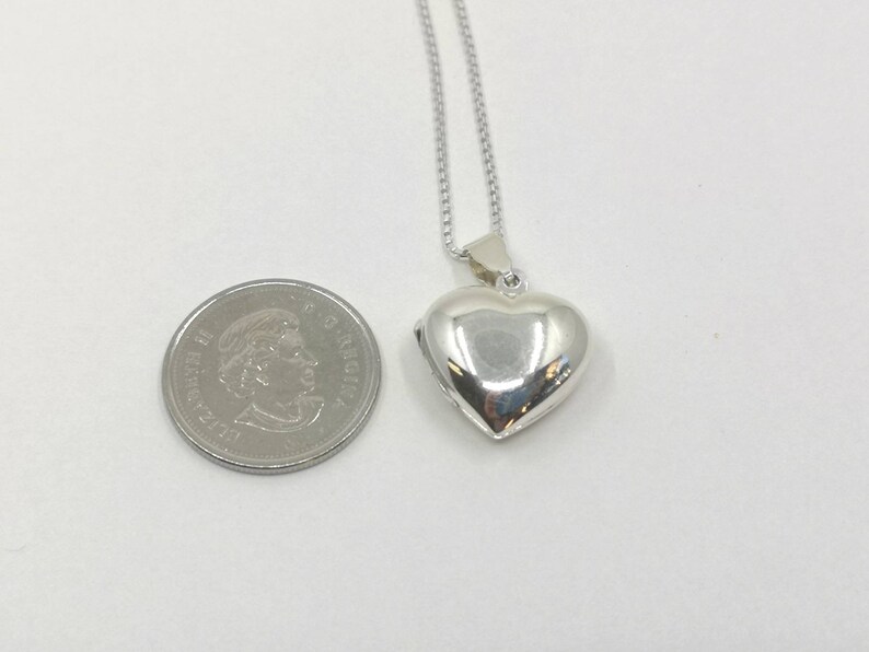 A sterling silver puffy heart locket on a silver chain. This heart necklace is on a white background next to a Canadian dime to show the size and scale of the pendant.