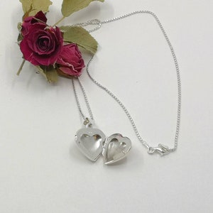 A sterling silver puffy heart locket on a silver chain. This heart necklace is on a white background with two dried red rose buds. The locket is shown open.