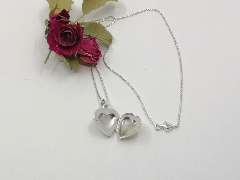 A sterling silver puffy heart locket on a silver chain. This heart necklace is on a white background with two dried red rose buds. The locket is shown open.