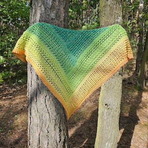 Op de afbeelding: Een handgemaakte, driehoekige sjaal in groen, geel en oranje, gedrapeerd over een boomstam. De sjaal heeft een open, kanten haakontwerp. De tekst "Szydełkowy Zakątek" is zichtbaar bovenaan.