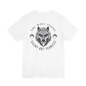 May include: White t-shirt featuring a detailed black and white illustration of a wolf's head. The design includes the text "THE WOLF INSIDE SILENT BUT FEARLESS" in a circular arrangement, with paw prints on either side.