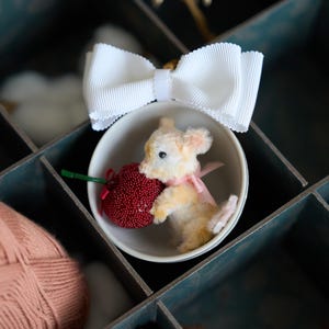Puede incluir: Una pequeña figura de ratón de peluche sosteniendo un corazón rojo con cuentas, metida en un cuenco blanco. Un gran lazo blanco se encuentra encima del cuenco. El ratón tiene una cinta rosa alrededor del cuello. La escena está en un compartimento color turquesa.