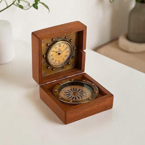 May include: A hinged wooden box containing a brass-toned analog clock with Roman numerals on the lid and a circular magnetic compass with the text Yay forge printed on the base. The clock face is gold-colored with black hands and markings.