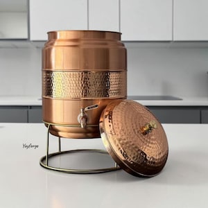 May include: A cylindrical copper water dispenser with a hammered texture band around the centre, a metal spigot, and a matching hammered lid resting beside it. The dispenser sits on a thin gold-toned wire stand. The text Yayforge is visible.