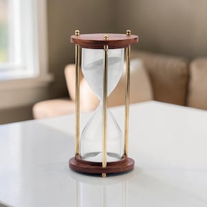 Puede incluir: Un reloj de arena con base y tapa de madera oscura, pilares de metal dorado y vidrio transparente con arena blanca. La pieza de madera superior tiene grabado el texto Yay forge. El dispositivo se encuentra en posición vertical.