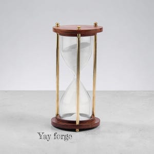 May include: An hourglass with a dark brown wooden top and base, connected by three vertical brass-colored metal rods. The clear glass bulb contains white sand. The text "Yay forge" is printed in a dark gray font near the base of the object.