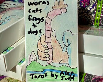 Könnte beinhalten: Tarotkarten-Deckbox mit einer skurrilen Illustration eines Wurms mit Hut, der von einer Hand gehalten wird. Der Text auf der Box lautet "worms cats frogs + dogs" und "Tarot by Alex Judd". Der Hintergrund ist hellblau mit einer grünen und blauen Landschaft.