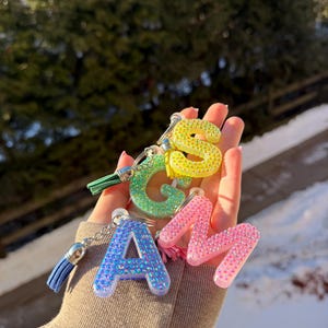 Pode incluir: Uma coleção de chaveiros iniciais coloridos. As letras A, M e S são visíveis, adornadas com strass brilhantes em azul, rosa e amarelo. Cada letra tem uma borla a condizer. A letra G é verde com uma borla verde.