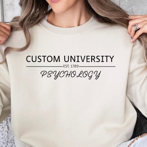 Könnte beinhalten: Cremefarbenes Sweatshirt mit dem Text "CUSTOM UNIVERSITY" über "PSYCHOLOGY". Das Sweatshirt hat auch den Text "EST. 1789" unter "CUSTOM UNIVERSITY". Das Sweatshirt ist eine neutrale Farbe und hat lange Ärmel.