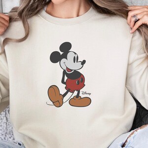 May include: A cream-coloured sweatshirt with a Mickey Mouse graphic. The cartoon character is in black, red, and brown, with the Disney logo. The long-sleeved top is a casual style.