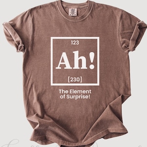 Peut inclure: T-shirt couleur expresso avec le texte "Ah!" dans un encadré de style tableau périodique, avec le texte "The Element of Surprise!" en dessous. Le t-shirt porte également le texte "Gulla Art Apparel" en bas.