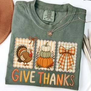 May include: A moss green t-shirt featuring a graphic with three rectangular frames containing a turkey, a stack of three pumpkins, and a plaid bow. The text "GIVE THANKS" is printed in orange and cream letters below the graphic designs.