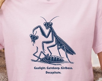 May include: A light pink t-shirt featuring a blue illustration of two praying mantises. The text below the illustration reads "Gaslight. Gatekeep. Girlboss. Decapitate.". The shirt is a casual, short-sleeved style.