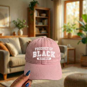 Pu&ograve; includere: Un cappellino da baseball rosa polveroso con la scritta bianca "PRODUCT OF BLACK HISTORY". Il cappellino ha una visiera curva ed &egrave; realizzato in un tessuto morbido. Il cappellino &egrave; mostrato davanti a un soggiorno.