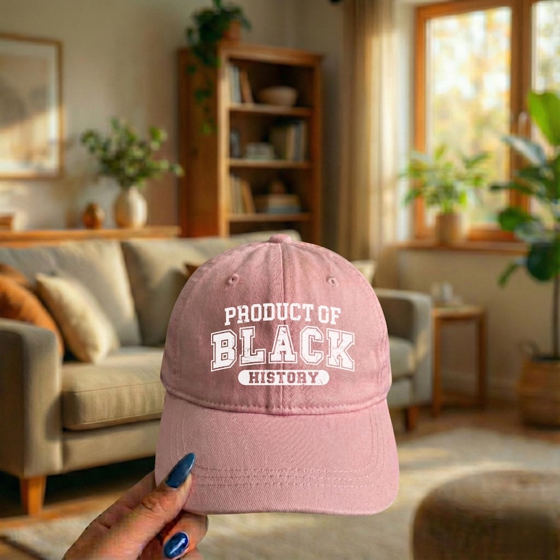 Pu&ograve; includere: Un cappellino da baseball rosa polveroso con la scritta bianca "PRODUCT OF BLACK HISTORY". Il cappellino ha una visiera curva ed &egrave; realizzato in un tessuto morbido. Il cappellino &egrave; mostrato davanti a un soggiorno.