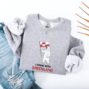 May include: A grey sweatshirt with a polar bear illustration holding a Greenland flag. The text "I STAND WITH GREENLAND" is printed in red. The sleeves are folded, revealing a white lining. The sweatshirt is made of a soft material.