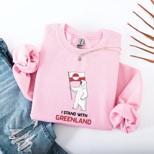 May include: A pale pink sweatshirt with a polar bear illustration holding a Greenland flag. The text "I STAND WITH GREENLAND" is printed in red. The sweatshirt is made of a soft material and is a crew neck style.
