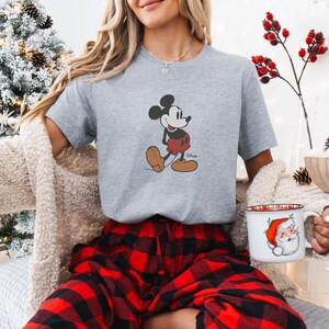 May include: A grey t-shirt with a Mickey Mouse graphic, worn with red and black plaid pyjama bottoms. A Christmas mug with Santa Claus is held. The background includes a decorated Christmas tree and festive decorations.