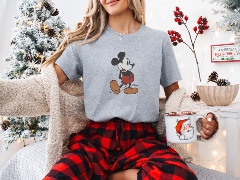 May include: A grey t-shirt with a Mickey Mouse graphic, worn with red and black plaid pyjama bottoms. A Christmas mug with Santa Claus is held. The background includes a decorated Christmas tree and festive decorations.