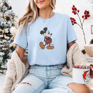 May include: Pale blue t-shirt with a Mickey Mouse design. The cartoon character is shown walking, wearing red shorts and brown shoes. This casual t-shirt is part of a clothing collection.