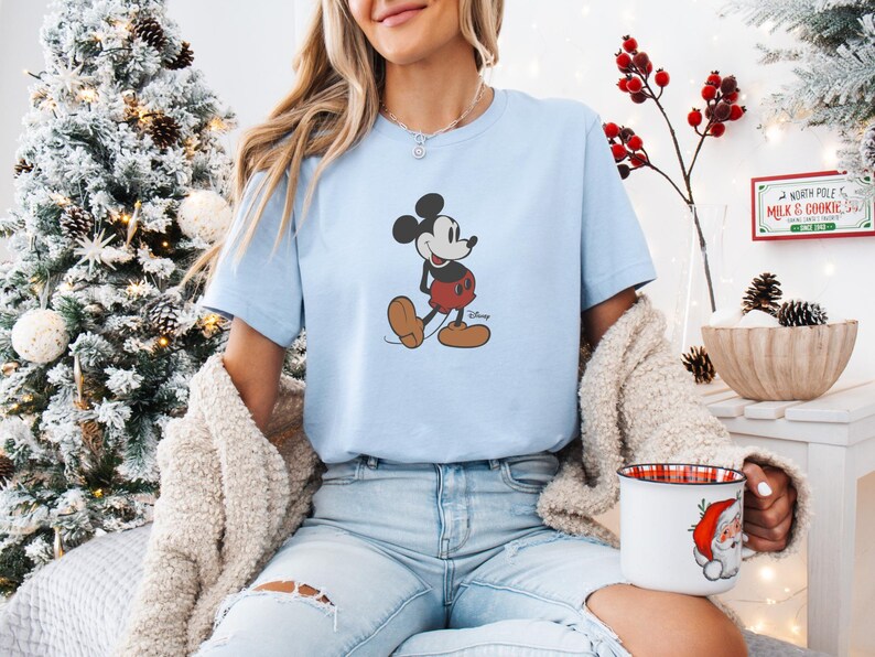 May include: Pale blue t-shirt with a Mickey Mouse design. The cartoon character is shown walking, wearing red shorts and brown shoes. This casual t-shirt is part of a clothing collection.